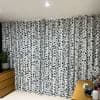 Full-length white fabric curtains with irregular black squiggle pattern, hanging from a ceiling track and drawn closed, filtering light; room corner with wooden dresser, potted plant, desk, ceiling light.