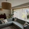 Sheer beige pleated curtains, drawn closed, filtering soft daylight over a corner living room with a gray tufted sectional, tall houseplants, pendant lamps, piano and a garden view beyond.