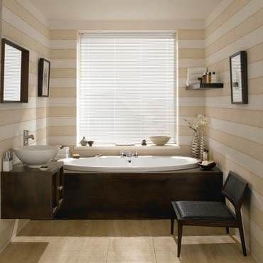 White horizontal Venetian blinds covering the central window, lowered and closed, filtering soft daylight; bathroom with dark wood-clad built-in bathtub, vessel sink, shelves, chair, striped beige walls and tiled floor.