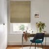 Roman fabric blind (olive‑yellow, leaf pattern), lowered, partially filtering daylight; above a desk with mirror, teal chair and vase of flowers in a neutral, softly lit room.