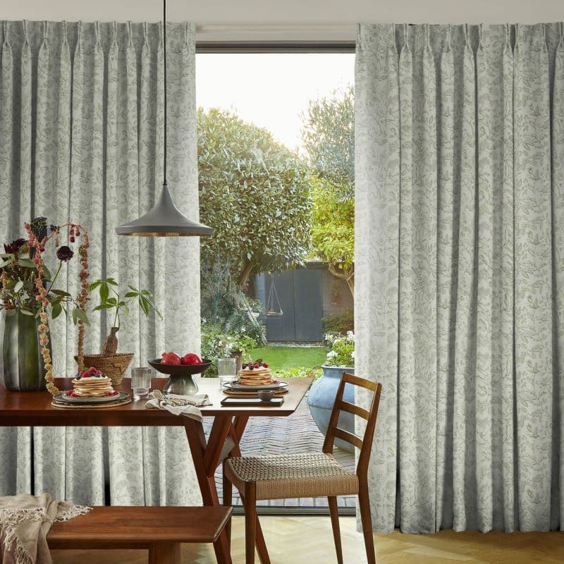 Floor-length pale curtains with subtle leaf print, drawn aside to frame a central open patio door, letting soft daylight into a dining area with wooden table set, chairs, garden beyond.
