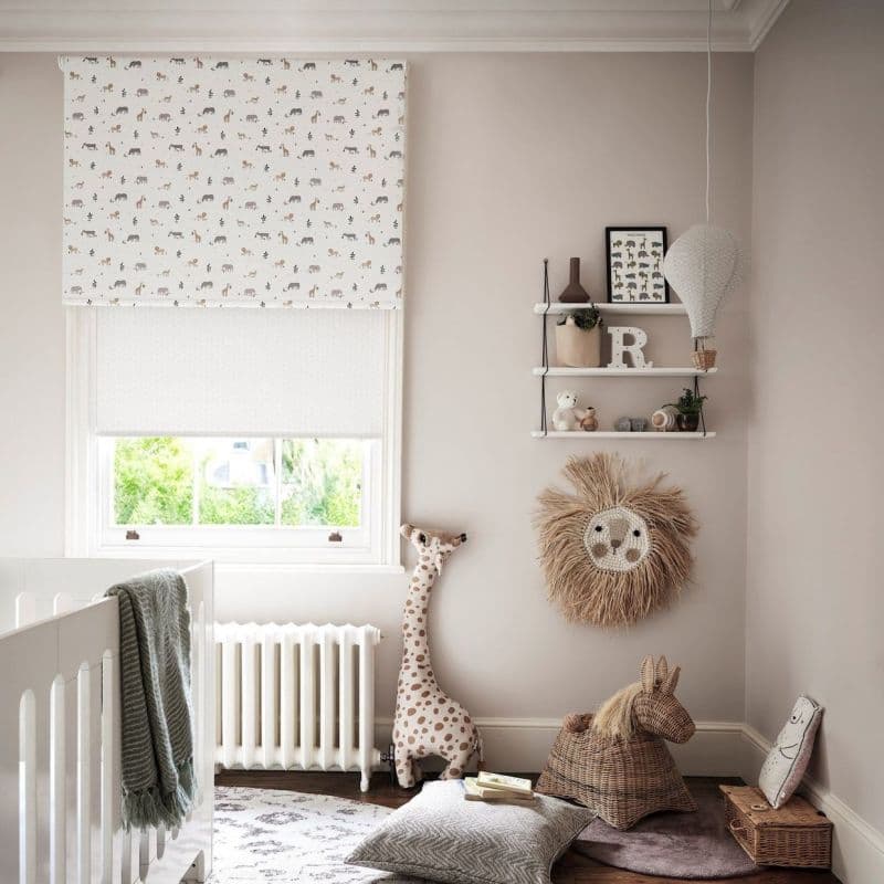 A white roller blind with small animal prints lowered over a window, filtering soft daylight; neutral nursery with crib, stuffed giraffe, wicker horse, shelves, hanging hot-air balloon and woven lion wall decor.
R