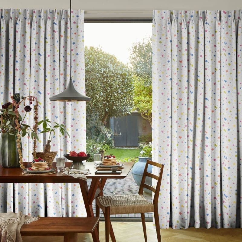 White pleated curtains with multicolored star print, pulled aside at center to frame a glass door and admit daylight; wooden dining table set for breakfast and leafy backyard beyond.