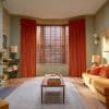 Rust-orange velvet curtains pulled aside revealing wooden Venetian blinds with tilted slats, filtering daylight in a bay window; surrounding cozy living room with sage walls, mustard sofa, wooden floors.