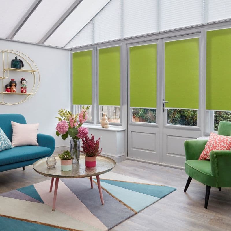 Green fabric roller blinds covering tall conservatory French doors and windows, lowered about three‑quarters; filtering daylight into a bright sunroom with a teal sofa, green armchair, coffee table, and flowers.