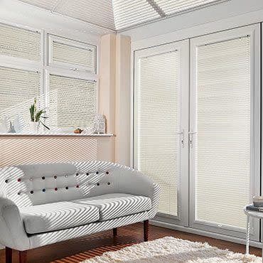 White pleated blinds fitted inside French doors and adjacent windows, light‑filtering; closed, softening daylight and casting striped shadows across a pale sofa and wood floor in a sunlit living room.