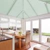 Pleated cellular blinds: pale mint on the pitched conservatory ceiling and white on windows and doors, closed and filtering daylight; bright kitchen-dining conservatory with wooden table, upholstered chairs, garden view.