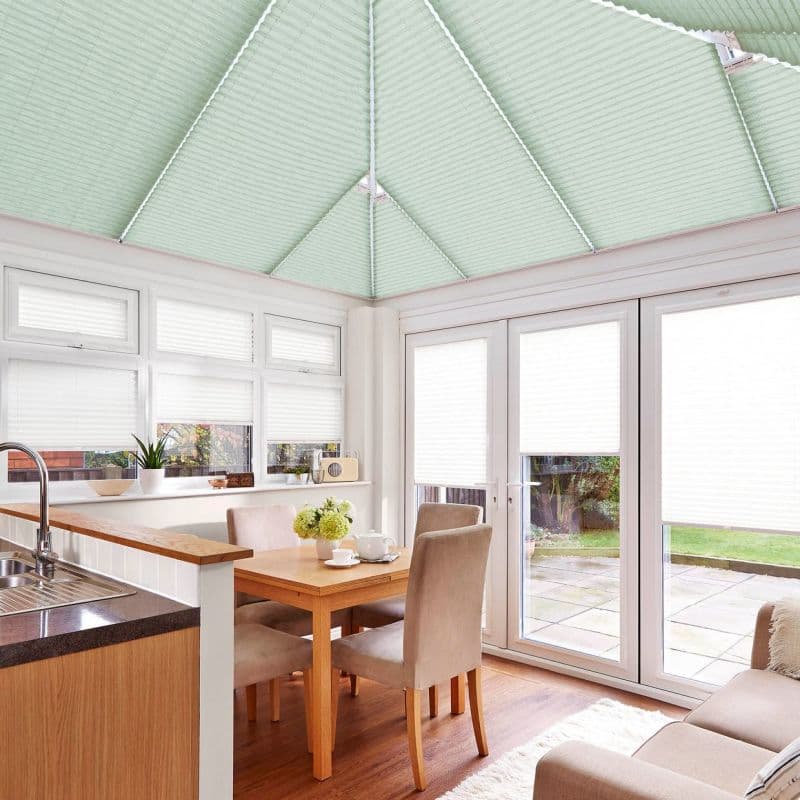 Pleated cellular blinds: pale mint on the pitched conservatory ceiling and white on windows and doors, closed and filtering daylight; bright kitchen-dining conservatory with wooden table, upholstered chairs, garden view.