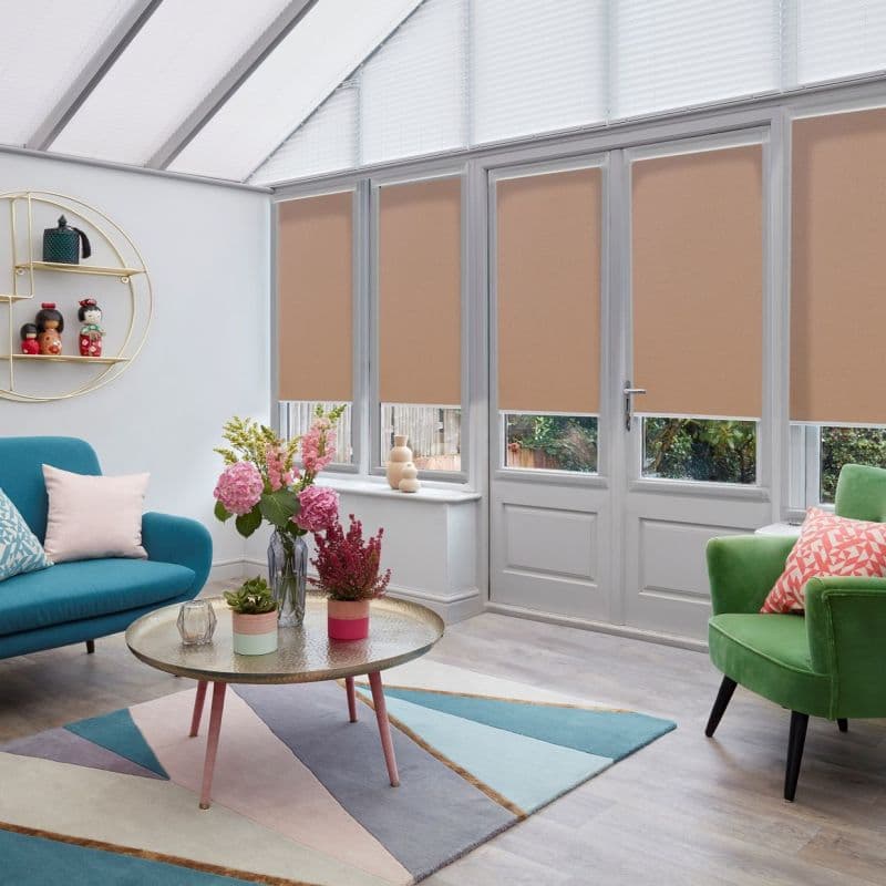 Beige roller blinds lowered mid-height over tall French doors and windows, filtering light; in a bright conservatory-style living room with glass roof, colorful sofas, geometric rug and flowered coffee table.