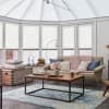 White cellular pleated blinds covering tall bay windows, lowered to mid-height and filtering soft daylight; bright conservatory-style living area with beige sofa, wooden coffee table and blue rug.