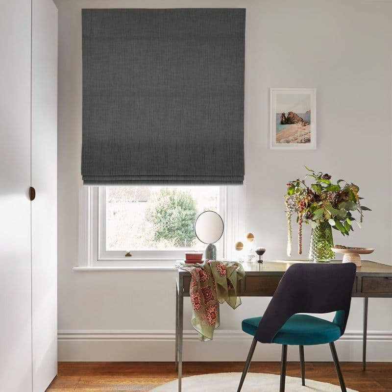 Dark grey textured Roman blind lowered, mostly closed and filtering daylight through a slim bottom gap; over a sash window above a desk with teal chair, vase of flowers.