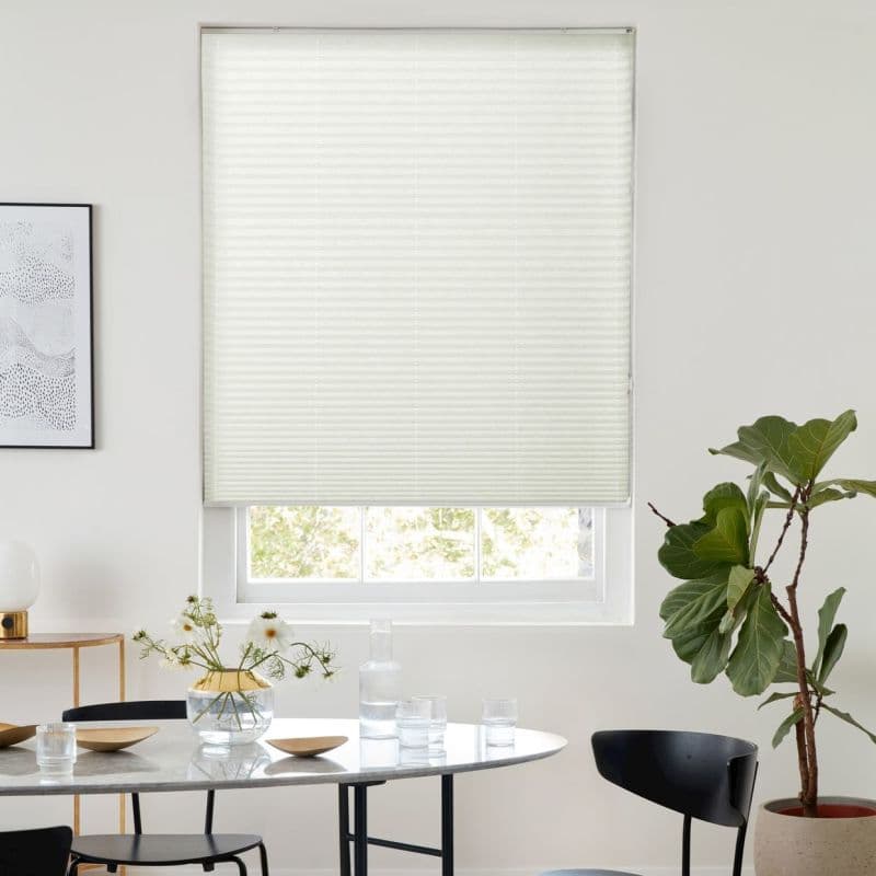 A white cellular pleated blind, lowered to cover most of the window and diffusing daylight; above a sunlit dining table with glassware, vase, black chairs and fiddle‑leaf fig.