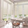 Object — Cream pleated blinds on tall windows and French doors. Action — Mostly lowered, filtering daylight through small gaps. Context — Bright dining nook with white table, chairs, floral cushions, garden view.