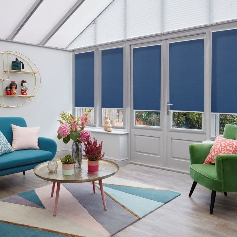 Navy roller blinds on four tall white-framed windows and a patio door, lowered over the lower two-thirds and filtering daylight, in a bright conservatory with colorful seating and a coffee table.