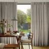 Beige full-length pleated curtains, gathered on each side and slightly parted at center revealing a sunlit garden; inside a dining table set for breakfast with wooden chairs and pendant lamp.