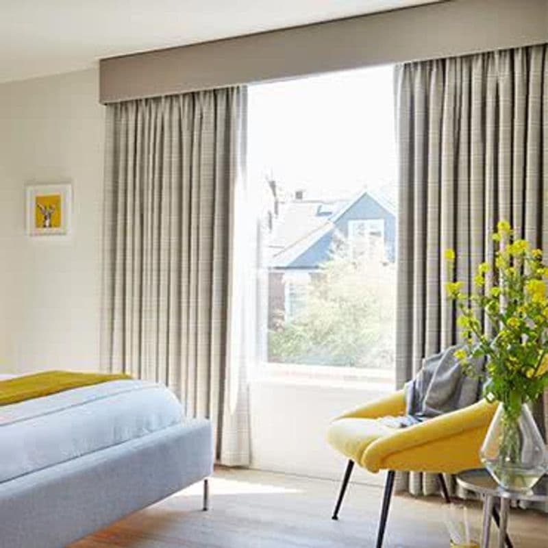 Floor-to-ceiling pleated beige drapes under a boxed valance, drawn to sides with a sheer opening — filtering bright daylight and slightly parted — modern bedroom interior with bed, yellow chair, vase, suburban view.