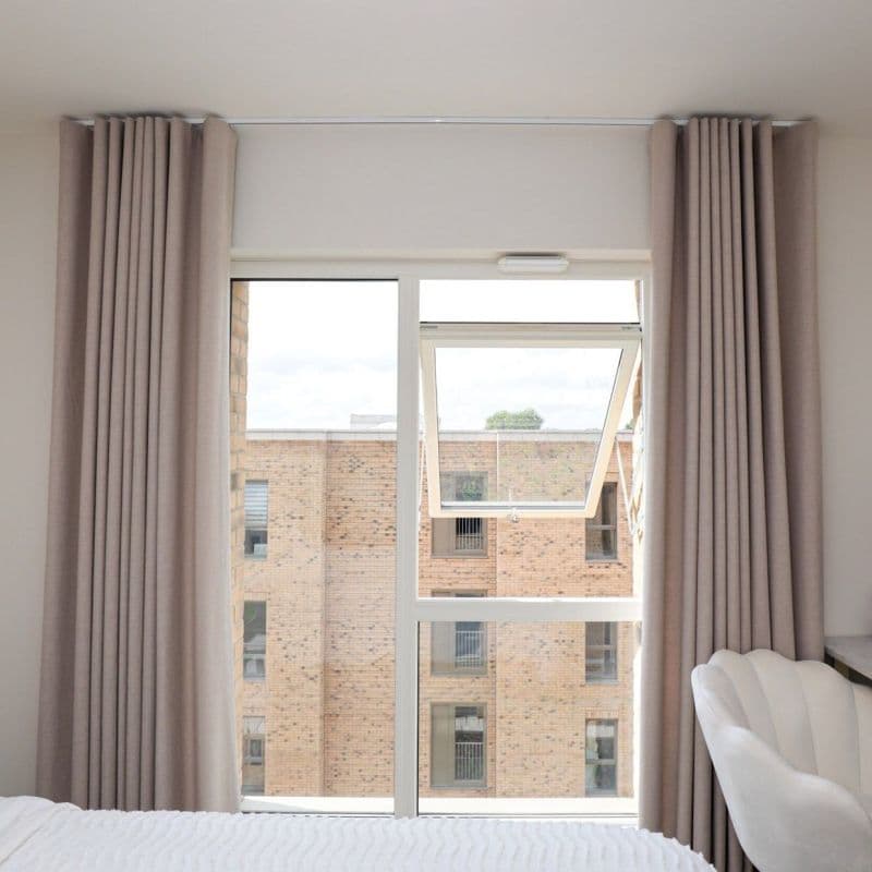 Floor-to-ceiling beige pleated curtains, pulled to each side, framing a white-framed window with its upper casement tilted open; bright bedroom interior, bed foreground, view of brick apartment building.