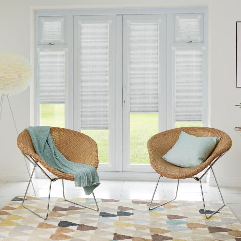 White internal Venetian blinds on tall French doors and sidelights, lowered and filtering daylight; set in bright minimalist living room with two wicker chairs, pastel throw, cushion, and patterned rug.