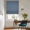 A textured blue Roman blind lowered almost fully, slightly folded at the bottom, filtering daylight through the window; a desk in a lit office with teal chair, vase of flowers.