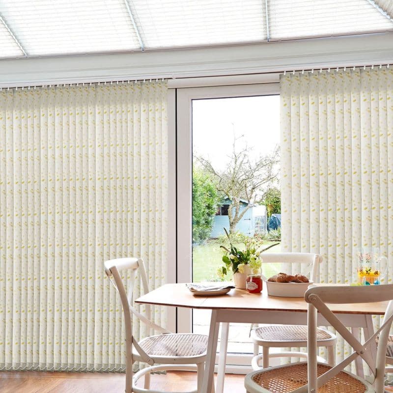 Vertical patterned fabric blinds cover a patio glass door, parted at the center and filtering soft daylight; in a bright dining/conservatory with wooden table, chairs, flowers, and a grassy backyard.