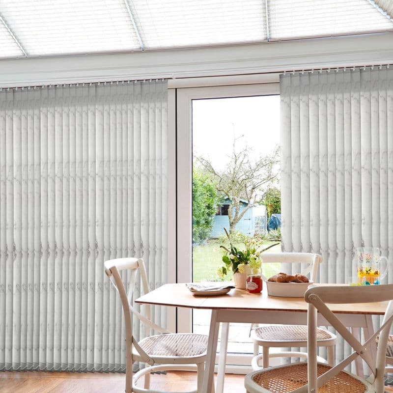 Pale patterned vertical blinds cover a full-height glass door, parted centrally to reveal the door; filtering daylight over a dining table with chairs, vase of flowers and a garden view.