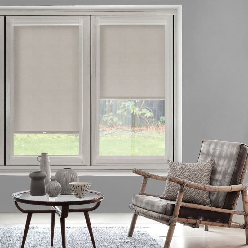 Beige fabric roller blinds covering twin windows, partially lowered and filtering daylight; mounted on windowsill in a grey living room with rattan armchair, small coffee table, and grassy backyard view.