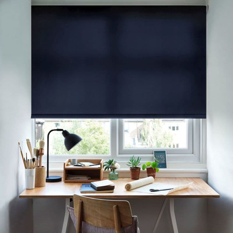 Dark navy roller blind, almost fully lowered, flat fabric across the window; blocking and dimming overhead light; above a wooden desk with lamp, plants and stationery in a bright home office.