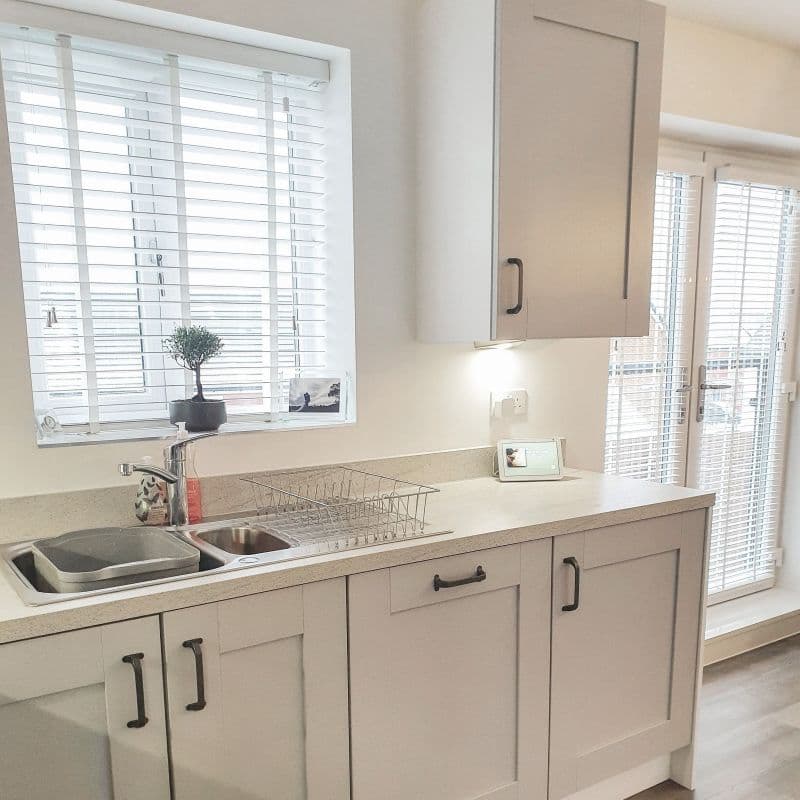 White horizontal blinds covering the sink window and adjacent French doors, slats tilted to filter bright daylight; pale kitchen with cabinets, stainless sink, dish rack, potted plant and smart display.