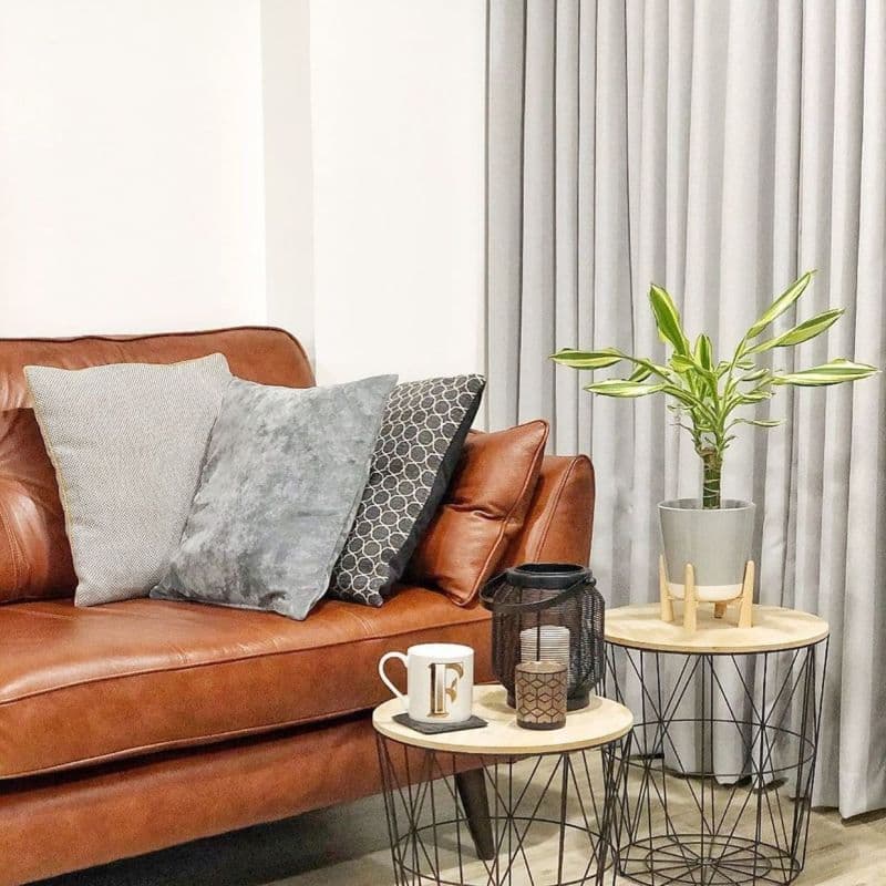 Grey floor-to-ceiling pleated curtains, drawn closed and softly diffusing light; living room with a brown leather sofa, three cushions, wire-frame nesting tables holding a potted plant, lantern, and mug. F