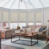 Cream pleated cellular shades, partially lowered across tall bay windows, filtering soft daylight into a bright conservatory-style living room with a light sofa, pink knit throw, wooden coffee table.