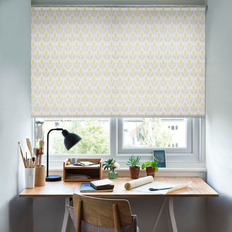 Patterned roller blind lowered over the window—pale grey fabric with yellow leaf motif—filtering soft daylight; tidy wooden desk beneath holds a lamp, art brushes, notebooks and small potted plants.