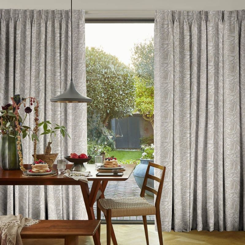 Full-length, pleated pale-grey curtains with leaf pattern, drawn open at center, framing a sunlit garden. Wooden dining table set for breakfast and pendant lamp in a cozy dining room.