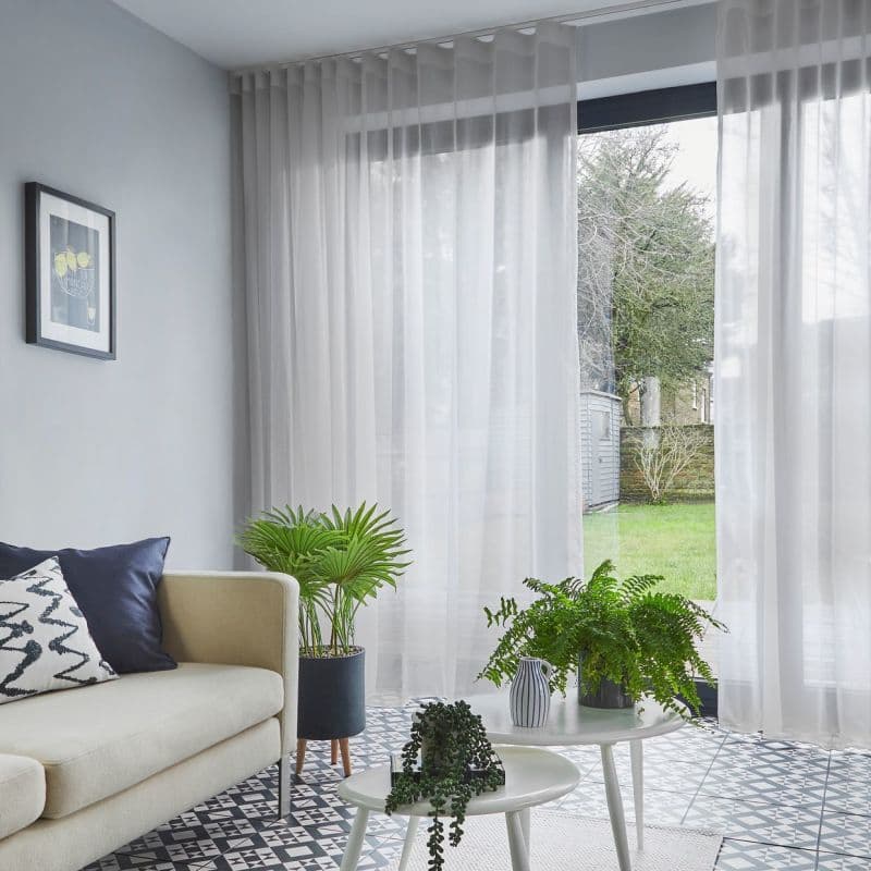 Sheer white, ceiling-mounted floor-to-ceiling curtains with pleats, partially drawn over sliding glass doors, filtering daylight into a living room with pale sofa, patterned tile floor, potted plants and lawn view.
