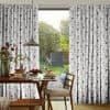 Pleated, floor-length curtains with gray–taupe geometric print, hung on a rod and parted centrally. They frame and soften sunlight through a sliding glass door. Dining table set for breakfast; leafy backyard beyond.
