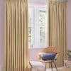 Beige gathered-pleat curtains (full-length, deep vertical folds) — mostly drawn, filtering soft daylight through a central white-framed window — pastel-pink-walled sitting corner with wicker chair, ottoman, and potted plant.