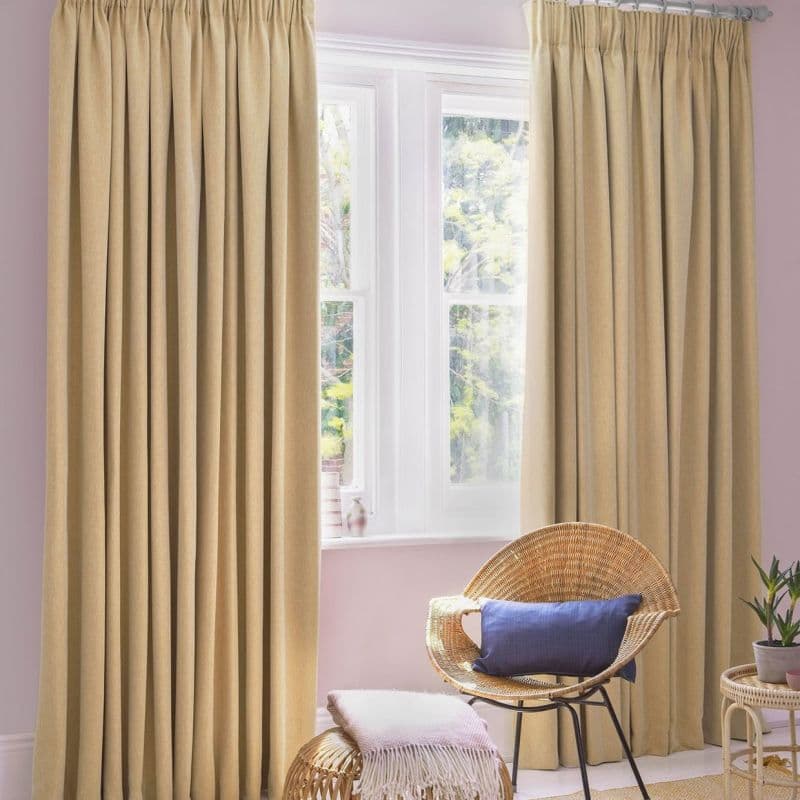 Beige gathered-pleat curtains (full-length, deep vertical folds) — mostly drawn, filtering soft daylight through a central white-framed window — pastel-pink-walled sitting corner with wicker chair, ottoman, and potted plant.