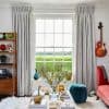 Floor-length pleated grey curtains, hung high and pulled to sides, framing a grid window admitting daylight; cozy living room with coffee table with vase, teal pouf, red sofa and guitar.
