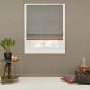 Gray fabric Roman shade with thin red trim lowered over a white-framed window, filtering soft daylight; taupe living-room wall, side table with potted plant and floor cushions nearby.