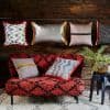 Decorative throw pillows—fringed, metallic, and patterned—hang from a horizontal rod and rest on a low red-and-black shag loveseat; rustic wood wall, cream patterned rug, black stool with potted fern.