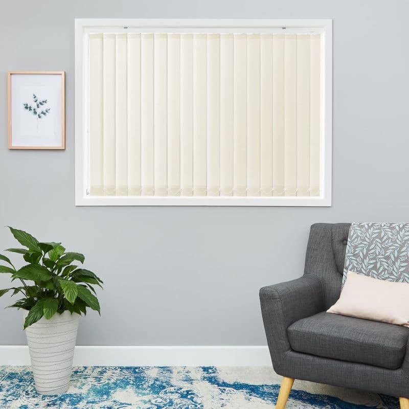 Cream vertical blinds covering a rectangular window, closed and evenly aligned, softly diffusing daylight; living room wall with gray armchair, patterned throw, potted plant and blue-accent rug.