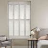 White plantation shutters covering a tall window—four framed panels with horizontal slats, closed and filtering soft daylight—set in a neutral bedroom corner with upholstered headboard, bedside lamp, vase, and armchair.