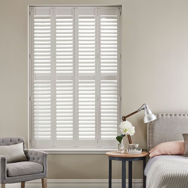 White plantation shutters covering a tall window—four framed panels with horizontal slats, closed and filtering soft daylight—set in a neutral bedroom corner with upholstered headboard, bedside lamp, vase, and armchair.