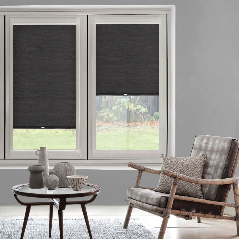 Two dark gray roller blinds on adjoining window panes, mostly lowered and filtering light; a gray living room with rattan-framed leather chair, patterned cushion, coffee table and backyard view.