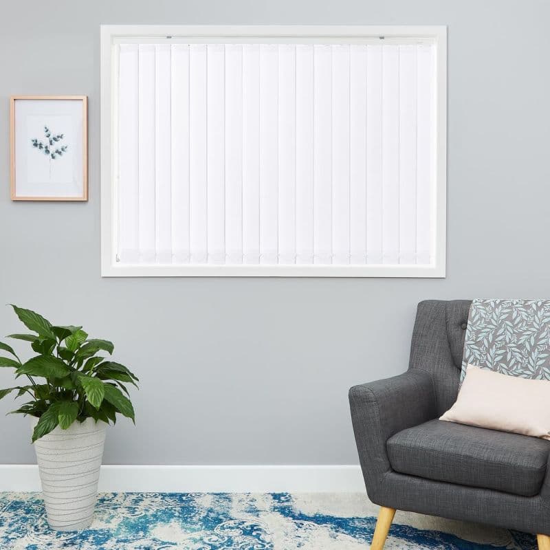 White vertical blinds inside the frame with evenly spaced slats, closed and softly diffusing daylight; in a grey-walled living room with grey armchair, patterned throw, potted plant and blue rug.