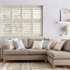 White plantation shutters covering a wide window, louvered slats tilted to filter soft daylight; set above a beige L-shaped sectional with pastel cushions in a bright, neutral living room.