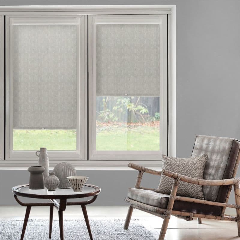 Two patterned roller blinds, lowered to cover most of a double window with translucent lower sections, filtering daylight; gray living room with rattan armchair, side table, decorative ceramics, and garden view.