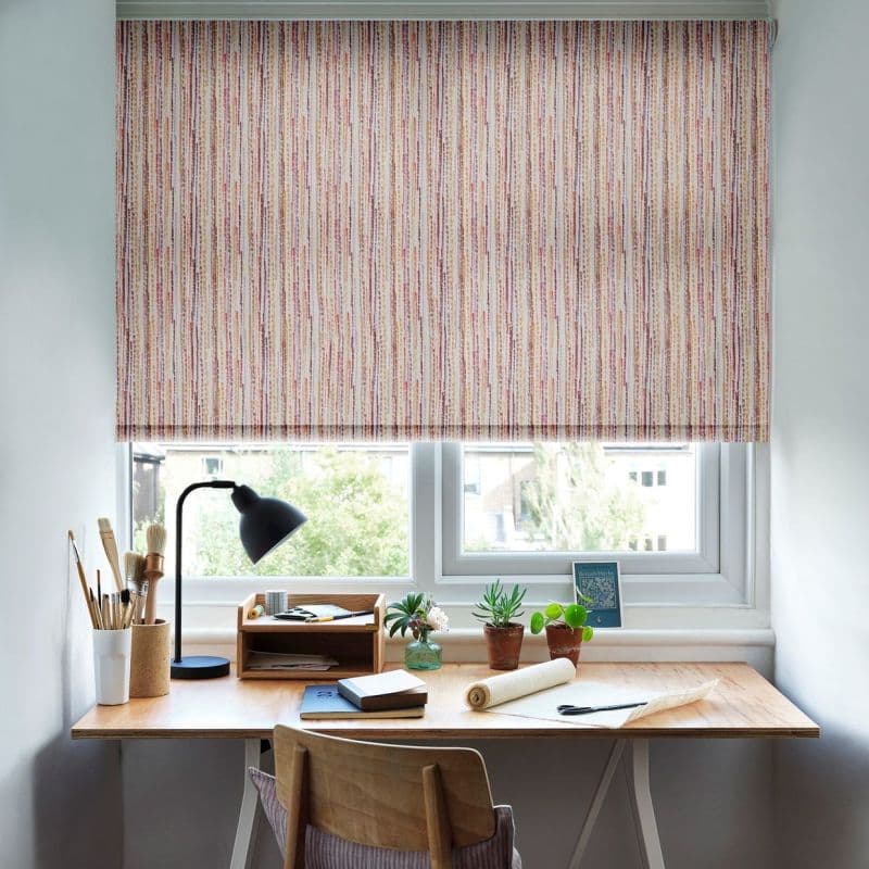 Roller blind with narrow vertical multicolored stripes, lowered partway, filtering daylight. Below, a wooden desk workspace with lamp, paintbrushes, notebooks, small potted plants; window reveals a leafy suburban view.