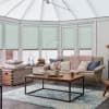 Mint-green cellular (pleated) blinds covering tall conservatory windows, lowered to just above the sills and filtering soft daylight into a cozy sunroom with a sofa, chunky knit throw, and wooden table.