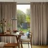 Beige pleated floor-length curtains, hung on a track and parted centrally, framing a sunlit backyard; wooden dining table set for breakfast with a pendant lamp in the foreground.