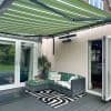 Green-and-white striped retractable awning overhead, fully extended and shading a patio; rattan L-shaped sofa with tropical cushions beside open folding doors on gray decking and a potted plant.
LOVE